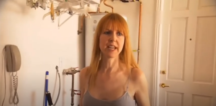una mujer rubia está en el cuarto de lavado de su casa lleva el cabello medio largo y con fleco trae una playera sin mangas gris 
