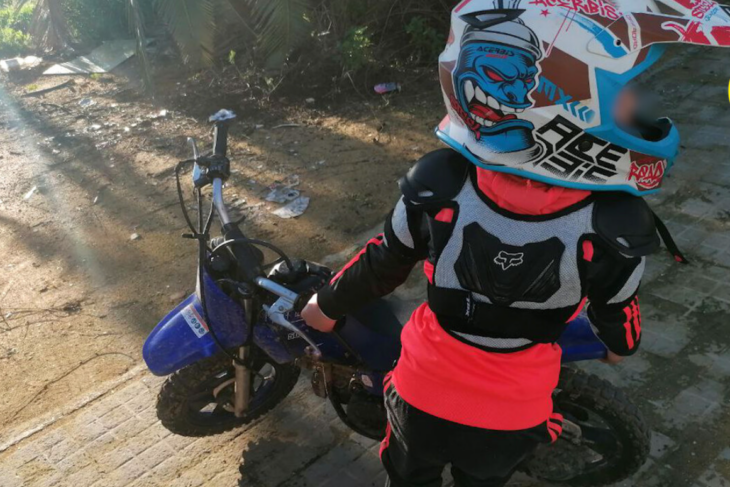 un niño de 7 años parado junto a su moto de color azul rey lleva casco y equipo de protección para moto en color naranja fluorescente 