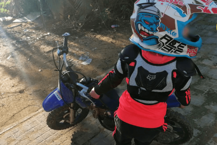 a 7 year old boy standing next to his royal blue motorbike is wearing a fluorescent orange motorbike helmet and protective gear 