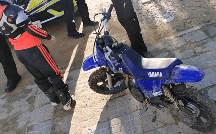 una pequeña moto de marca Yamaha en color azul rey con llantas negras se observan otras personas cerca del vehículo que está estacionado sobre una calle