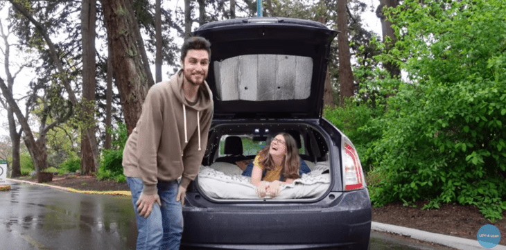 a couple smiles at the camera the woman is lying inside a black car with the trunk open he is standing outside the car