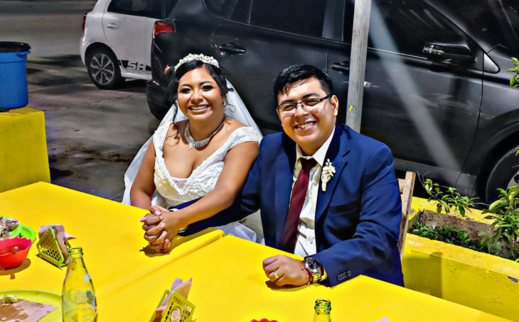 some bride and groom in their wedding suits sat at the table of a street taco stand they are holding hands and smiling happily 