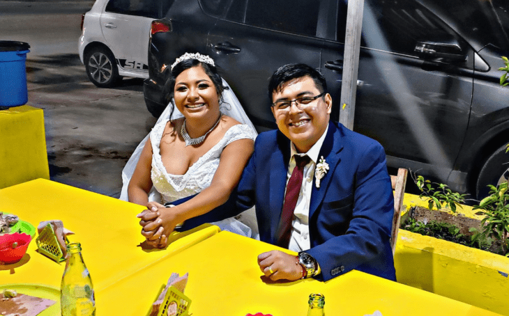 unos novios con sus trajes de boda se sentaron en la mesa de un puesto callejero de tacos están tomados de la mano y sonríen felices 