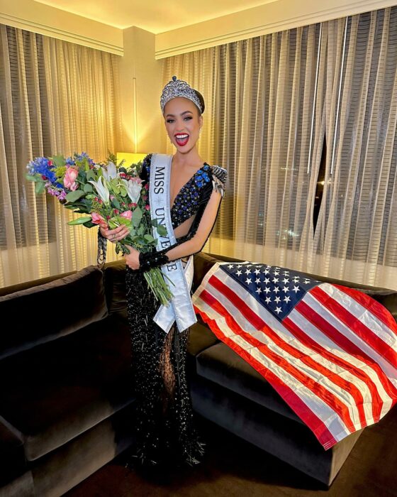 R´Bonney Gabriel la Miss Universo USA con flores y una bandera de Estados Unidos en una sala 