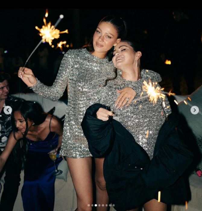 Selena Gomez y Nicola Peltz recibiendo el año nuevo traen en la mano una luz de bengala llevam vestidos de Valentino en tono gris plata y están recargadas una en la otra como señal de amistad Selena lleva una chamarra negra semi puesta 