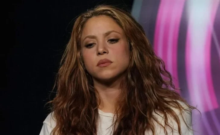 la cantante Shakira luce la mirada baja y lleva el cabello suelto y alborotado con chinos su maquillaje es marcado trae una blusa de color claro 