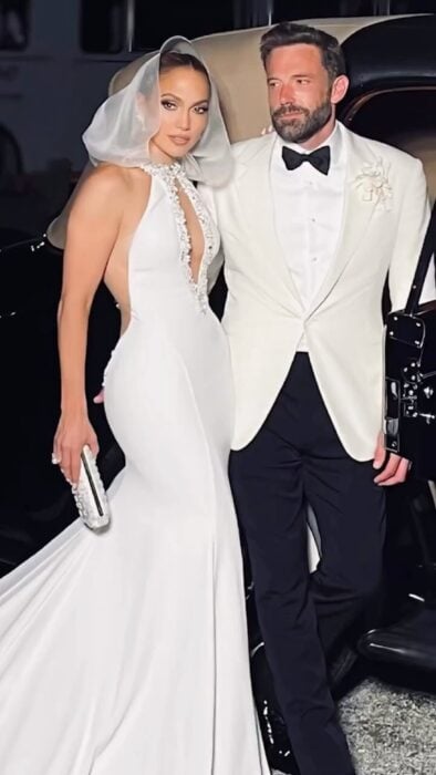 JLo junto a Ben Affleck en el día de su boda ella está vestida de novia con un traje blanco y velo en la cabeza él lleva un esmoquin blanco con moño negro