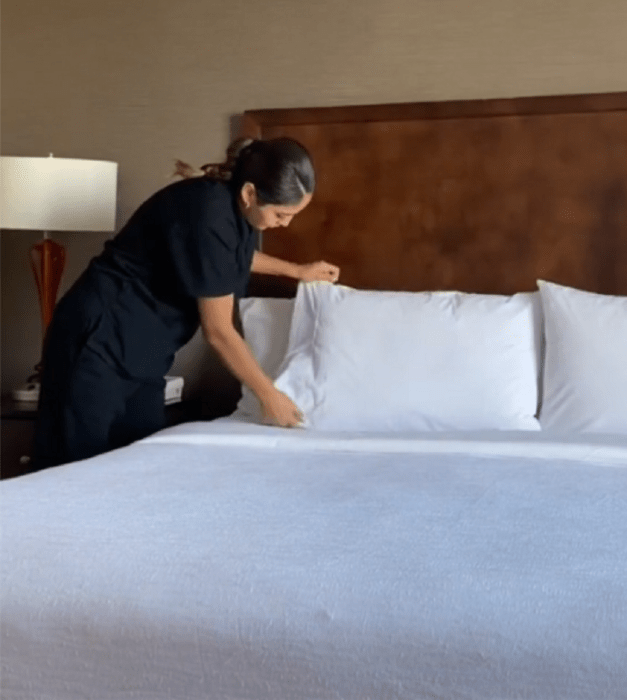 la empleada de limpieza de un hotel acomodando las almohadas de una cama con sabanas blancas 