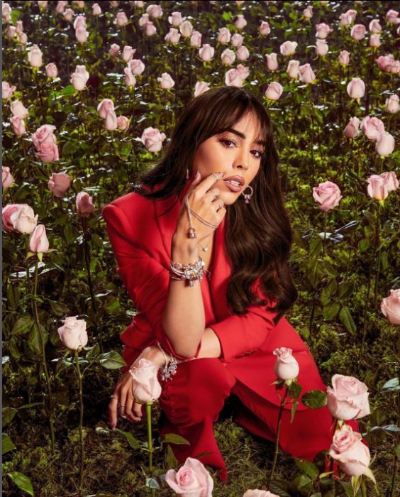 Danna Paola vestida de rojo en cuclillas sobre el pasto de un jardín rodeada de muchas rosas rosas