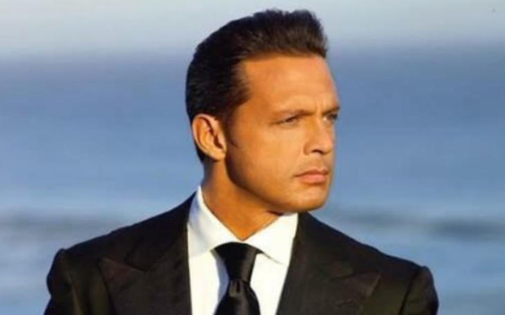 Luis Miguel posando vestido con un traje y corbata negros y una camisa blanca de fondo se ve el mar