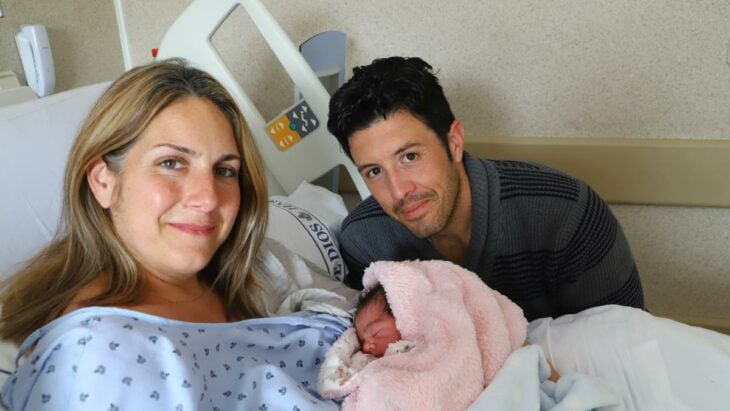 papá y mamá en un cuarto de hospital con su bebita recién nacida, su madre la sostiene en brazos mientras está acostada en la cama