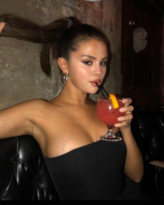Selena Gomez tomando una bebida en una copa usando un atuendo en color negro 