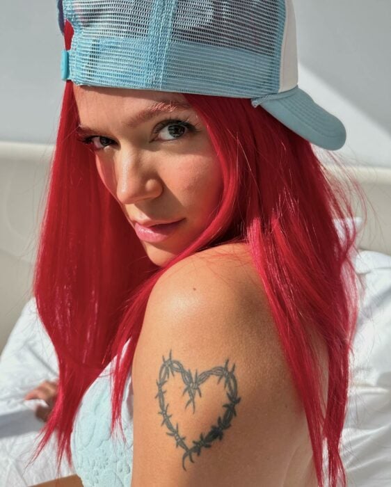 Fotografía de Karol G con una gorra color verde mostrando su tatuaje de corazón 