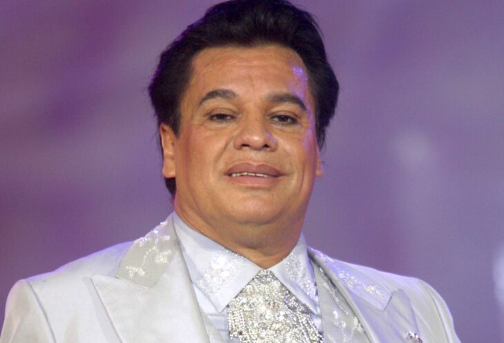 primer plano del cantante fallecido Juan Gabriel lleva un traje blanco con pedrería el fondo de la imagen es de color morado