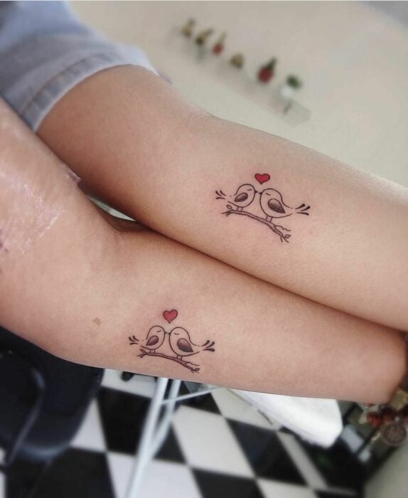 dos personas mostrando un tatuaje con figura de pajaritos dándose un beso con un corazón en medio 