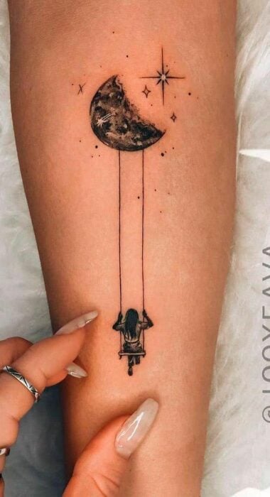 brazo de una persona mostrando el tatuaje de una luna con una mujer columpiándose en ella 