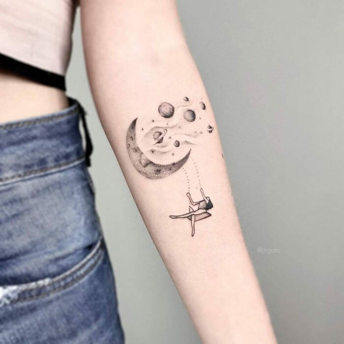 brazo de una chica mostrando el diseño del tatuaje de una luna con unos planetas mientras se columpia en ellos 