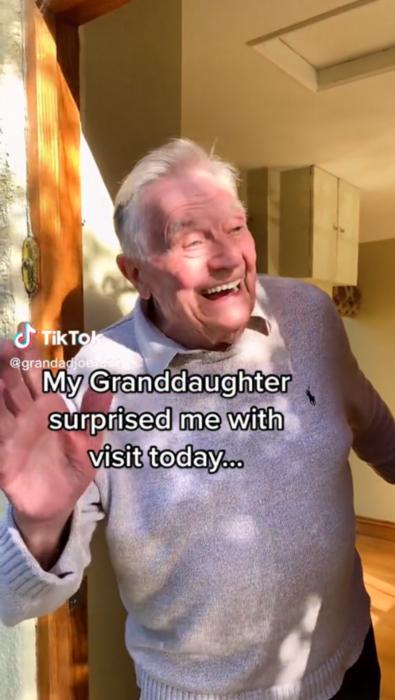 un abuelito sonríe a la cámara mientras saluda con su mano derecha
