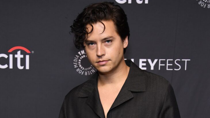 Cole Sprouse. posando en un evento lleva una camisa oscura y el cabello un poco largo del frente