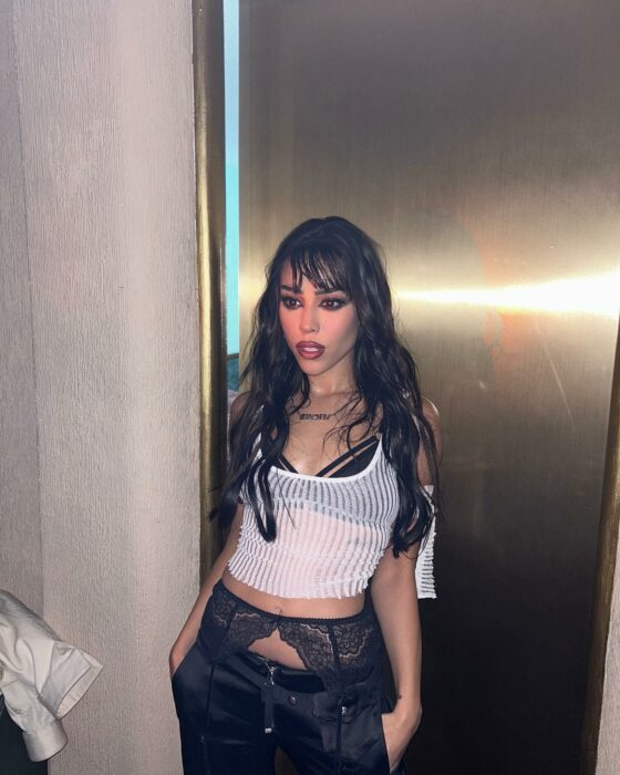 Danna Paola posando con una blusa transparente blanca recargada en una puerta 