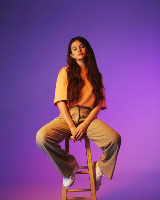 Selena Gomez posando frente a un fondo morado sentada en un banco 