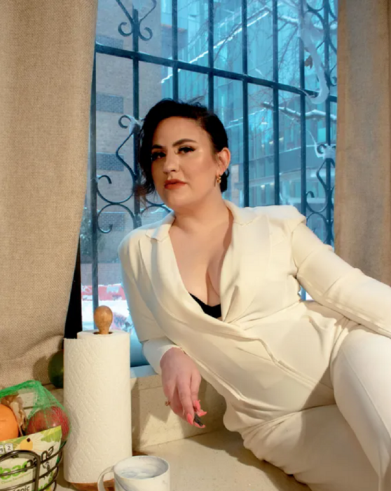una mujer lleva un traje blanco sastre y está recargada sobre un mueble de la cocina tras de ella hay una ventana