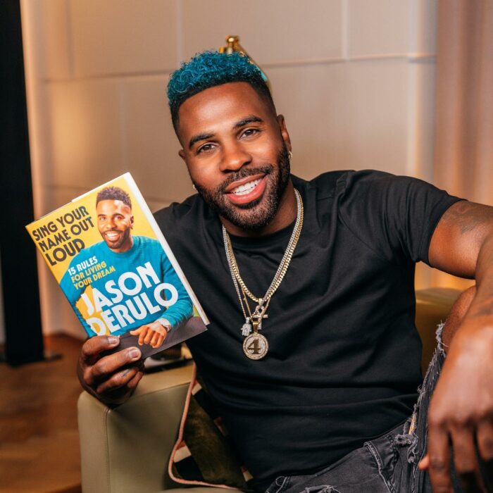 Jason Derulo sentado con un libro en su mano mostrándolo a la cámara