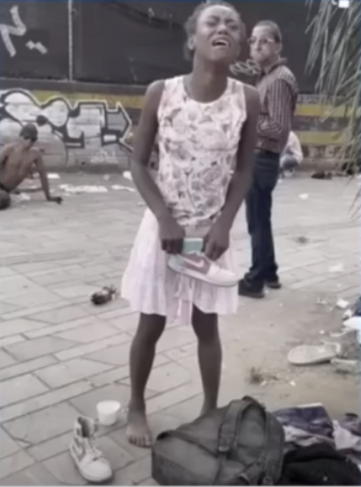 una chica afroamericana en situación de calle está descalza y sostiene un zapato teni en sus manos