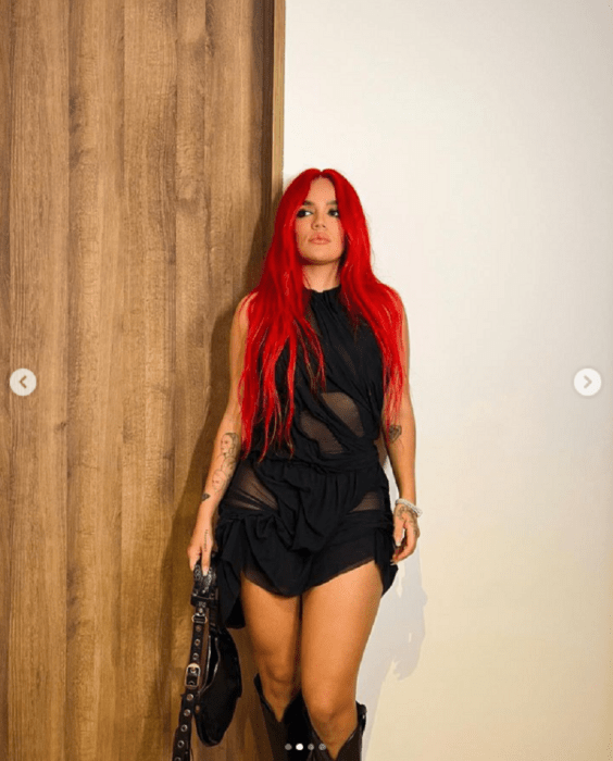 Karol G posando con un vestidito negro lleva el cabello largo y rojo está parada junto a una puerta