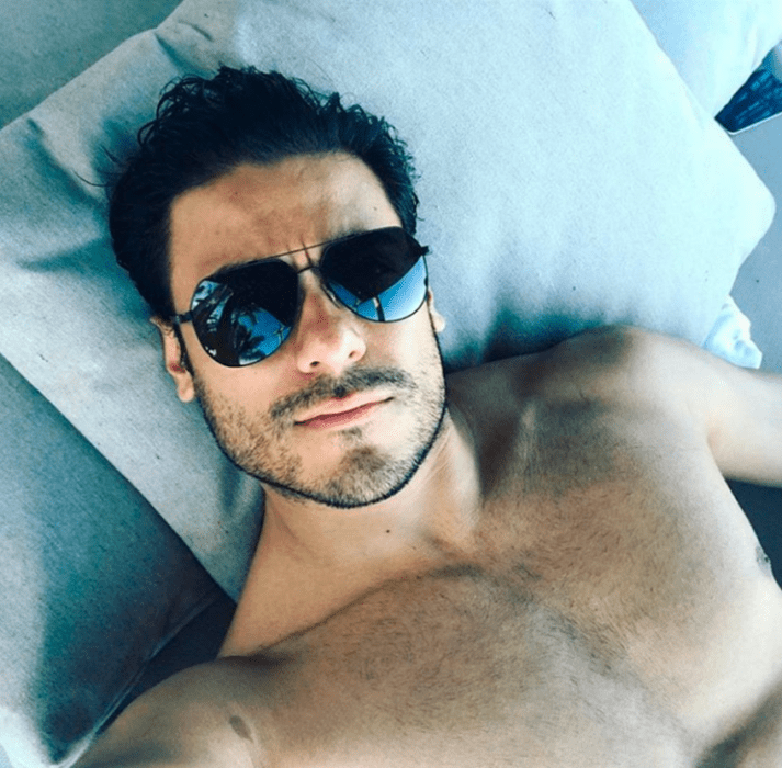 Carlos Rivera con lentes oscuros y el torso desnudo acostado en una cama