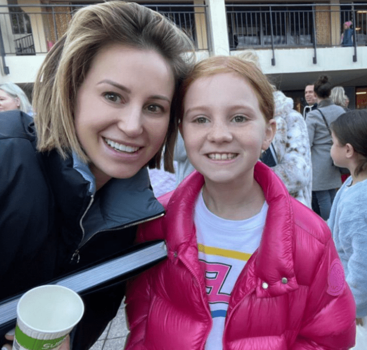 a caucasian mother and daughter smile at the camera they are both wearing casual clothes and are on the street