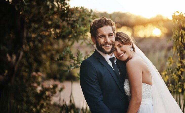 unos novios posando sonrientes en una jardín al atardecer ella se recarga cariñosamente en el hombro de él