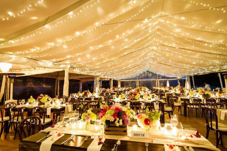 una boda llena de luces cálidas en el techo en las mesas hay adornos de flores y velas no hay invitados 