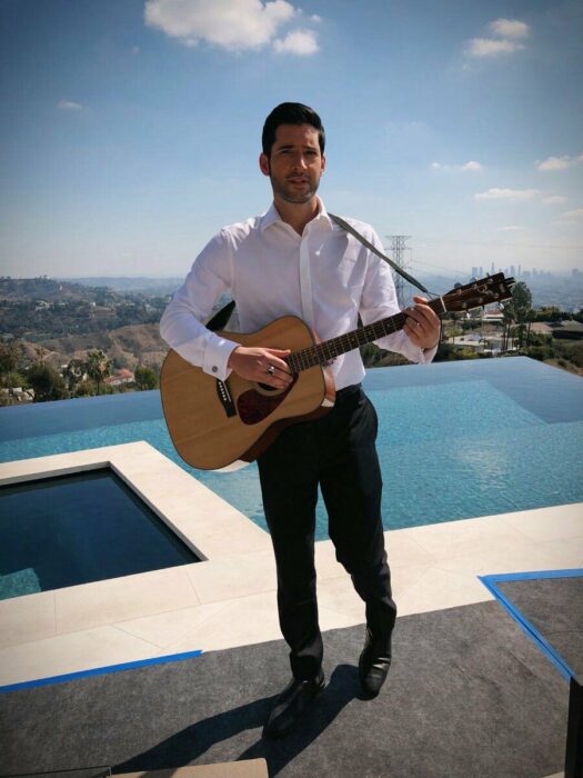 Tom Ellis tocando la guitarra a la orilla de de una alberca 