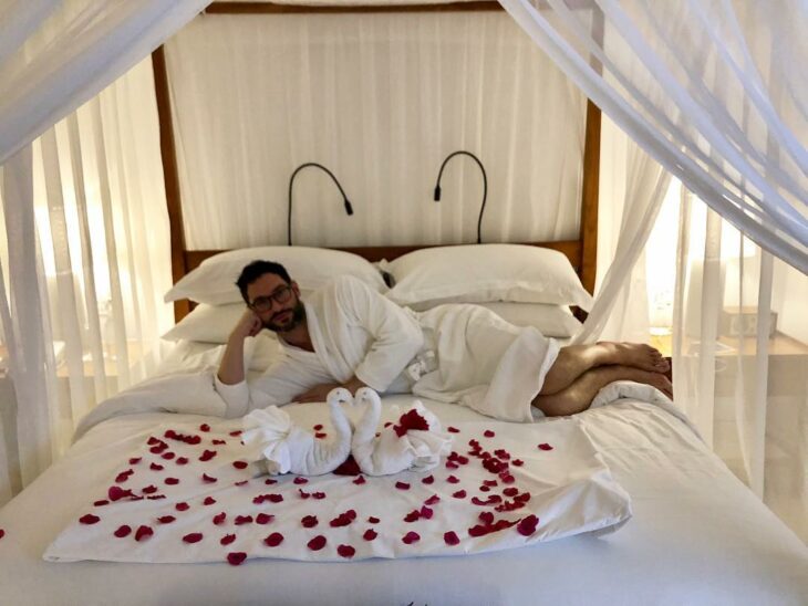 Tom Ellis acostado en una cama con algunos pétalos de rosas rojas 