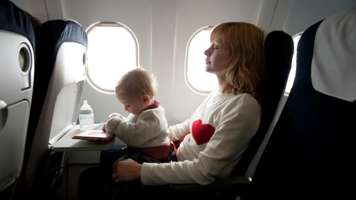una mamá de un bebé descansa en el asiento de un avión con su bebé en sus piernas la bebé juega con un pequeño libro de dibujos