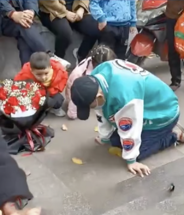un hombre arrodillado en el pavimento con un ramo de flores a un lado gente lo observa 