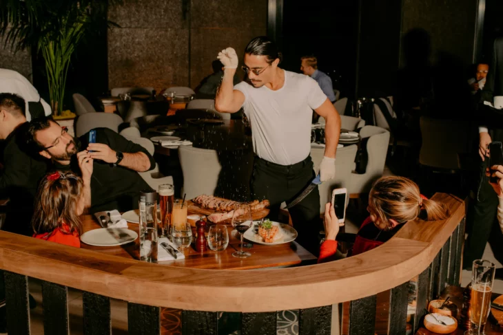 el chef Salt Bae haciendo su movimiento característico al ponerle sal a los alimentos un grupo de clientes de su restaurante lo observa