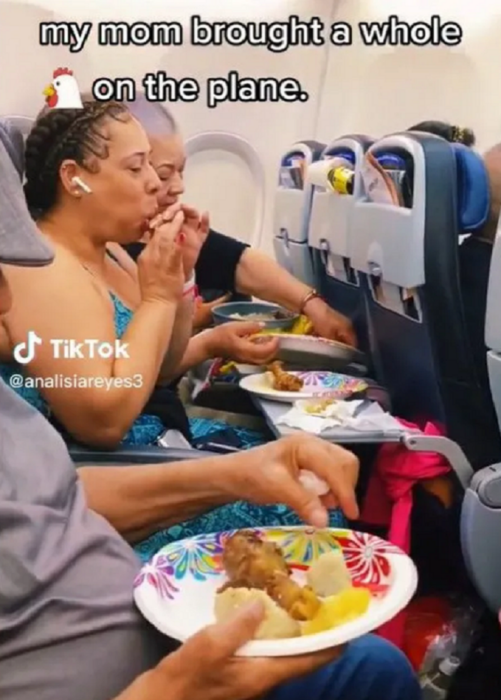 una mujer come con las manos una pieza de pollo frito mientras está sentada en el asiento de un avión las personas de su fila también tienen comida en platos desechables