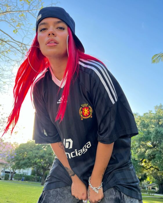 KArol G posando con camisa de Fútbol y gorra en un jardín 