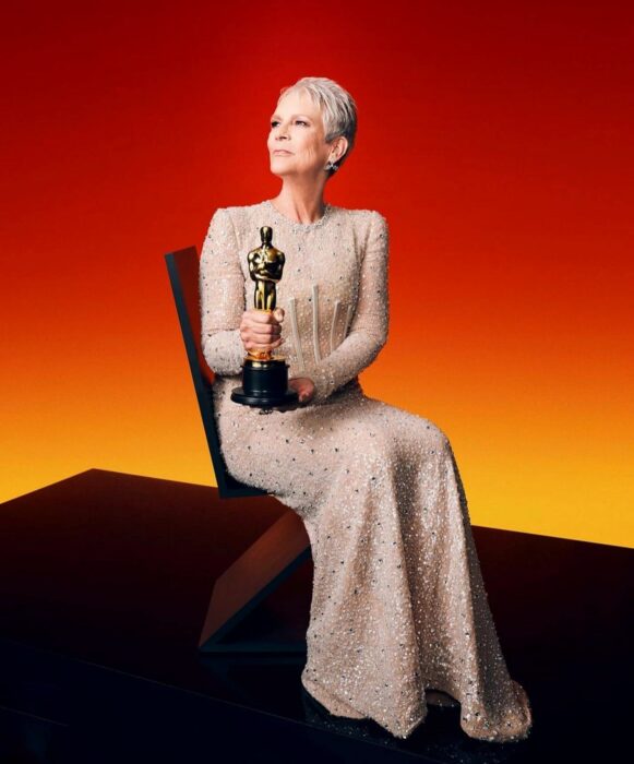 Actriz Jamie Lee Curtis posando con su galardón Oscar 2023 en un fondo color rojo con amarillo 