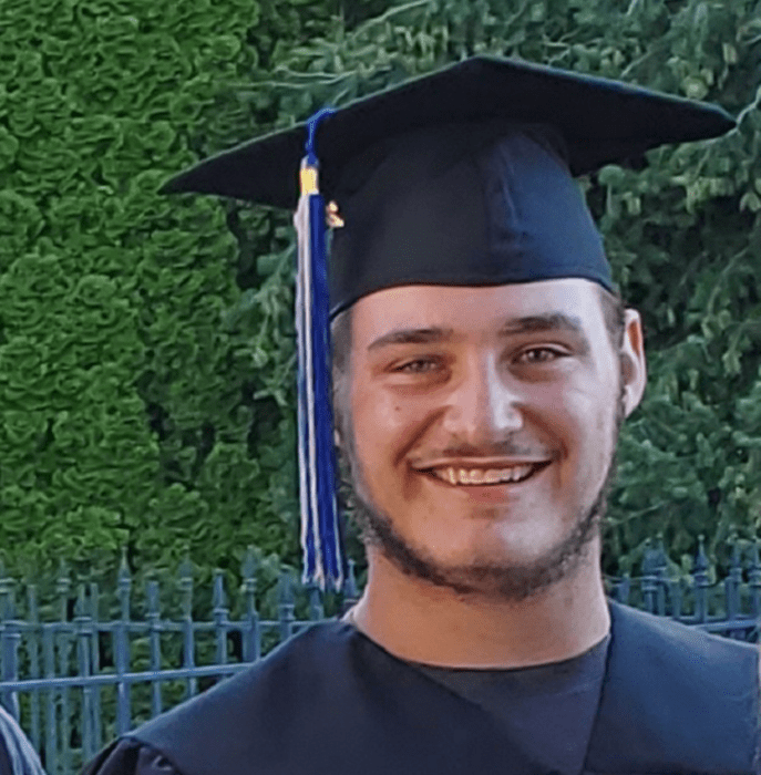 un joven graduado posa sonriente vestido con toga y birrete en color azul marino 