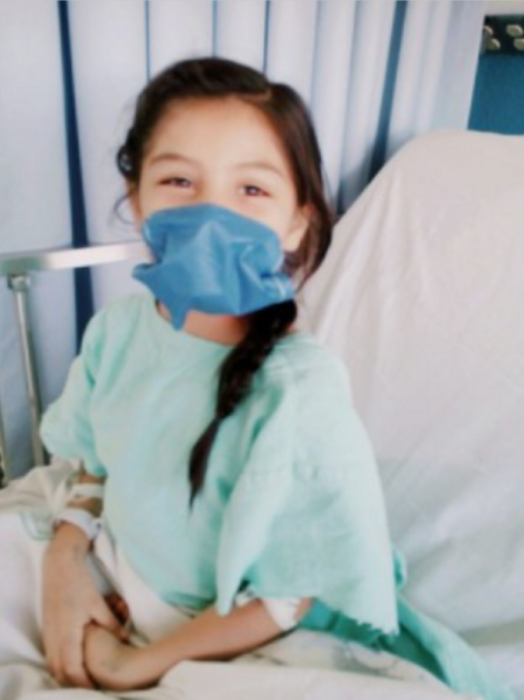 una niña está en una cama de hospital lleva en el rostro un cubrebocas azul y una bata verde esmeralda 