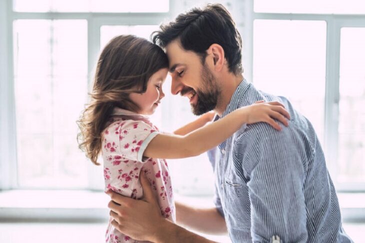 un papá junto a su hija están uniendo sus frentes y la niña toca los hombros de su papá, y el la sostiene de la cintura