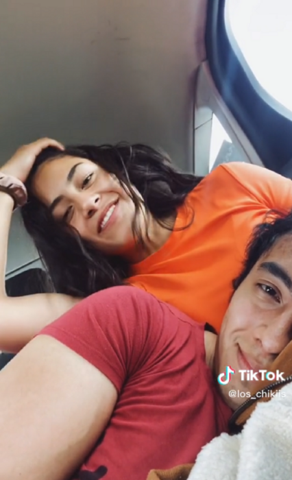 una pareja sonríe a la cámara mientras están en una auto él está recargado acostado sobre las piernas de ella