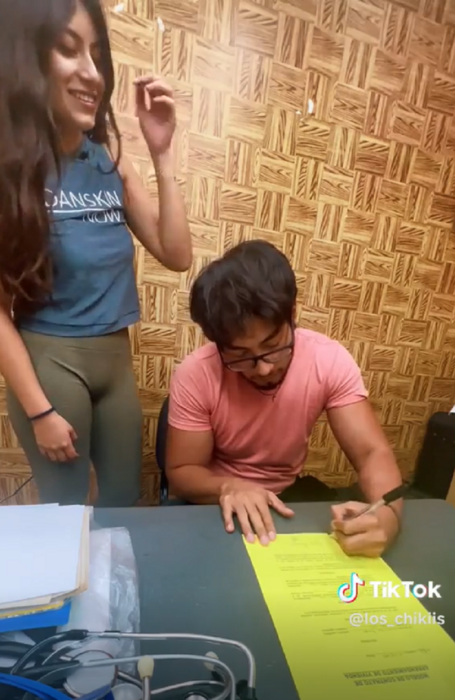 un hombre firma un contrato mientras una chica está de pie a su lado están en un cuarto que parece ser una oficina