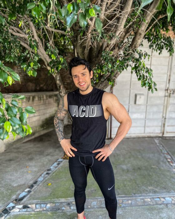 Fotografía de un chico posando frente a un árbol en el patio de un lugar con un atuendo en color negro 