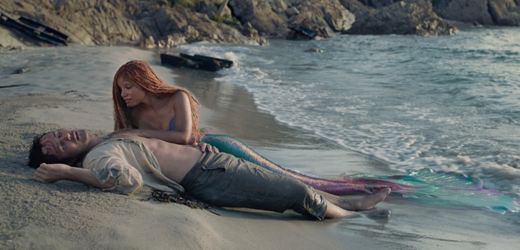 una imagen de la nueva película de La Sirenita live action Ariel y Eric están en la playa el príncipe está inconsciente acostado en la arena