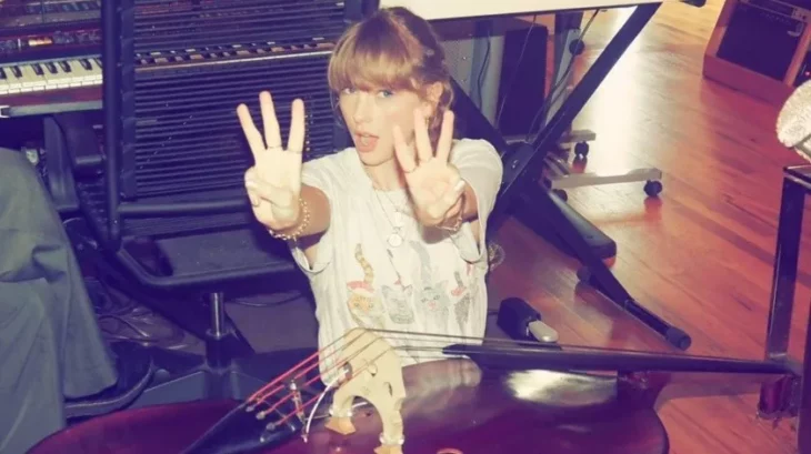 Taylor Swift muestra con sus dedos un tres en cada mano, está junto a un piano