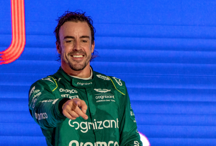 El piloto de F1 Fernando Alonso sonríe mientras señala a alguien en el público lleva su traje verde de corredor de autos 